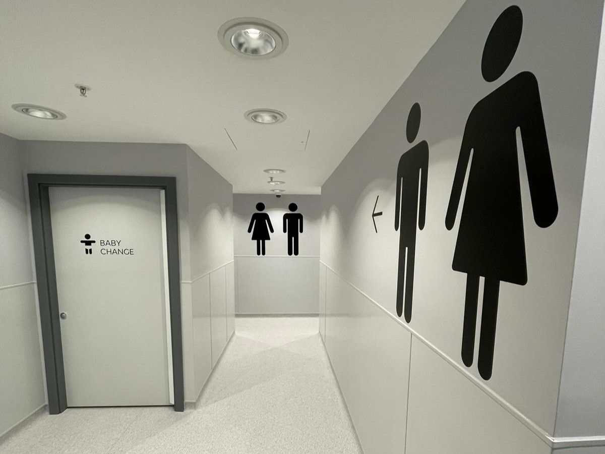 Public restroom corridor with grey and white hygienic wall cladding panels, gender signage, and baby change room door