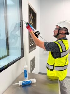 Technician applying Chem Link DuraSil sealant around a utility panel during hygienic wall cladding installation in a cleanroom environment